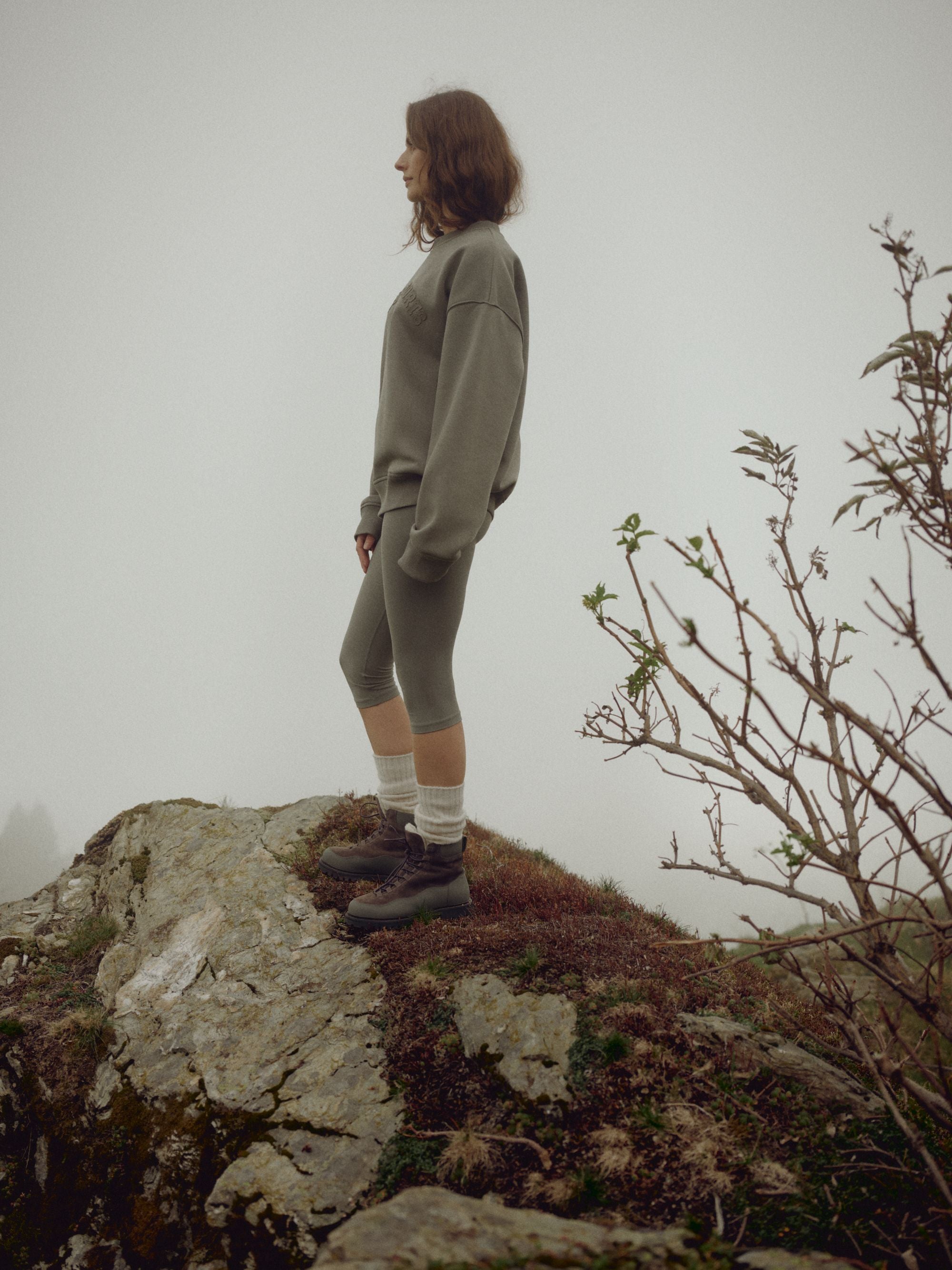 Woman wearing a khaki CASPARA CS crewneck sweatshirt standing on a rocky outcrop in the fog.